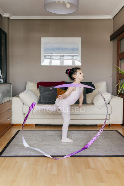 Vista laterale di carina bambina in body e collant nastro rotante e ballare durante la formazione ginnica ritmica in accogliente soggiorno a casa — Foto stock