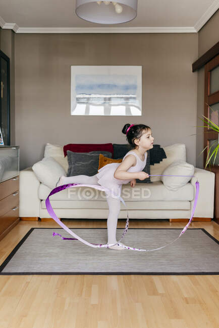 Nettes kleines Mädchen in Trikot und Strumpfhose, das Band dreht und beim rhythmischen Gymnastiktraining im gemütlichen Wohnzimmer zu Hause tanzt — Stockfoto