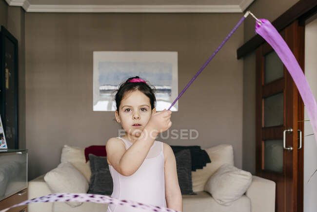 Focused cute little brunette girl in leotard looking at camera while spinning ribbon during rhythmic gymnastic practice training in cozy living room at home — Stock Photo
