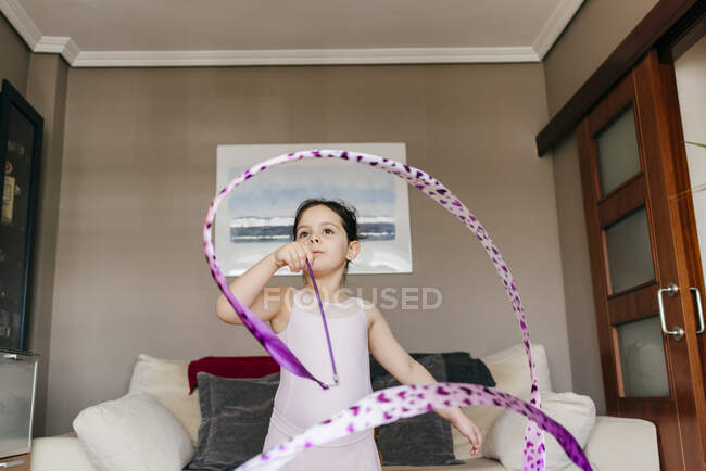 Focused cute little brunette girl in leotard looking away while spinning ribbon during rhythmic gymnastic practice training in cozy living room at home — Stock Photo