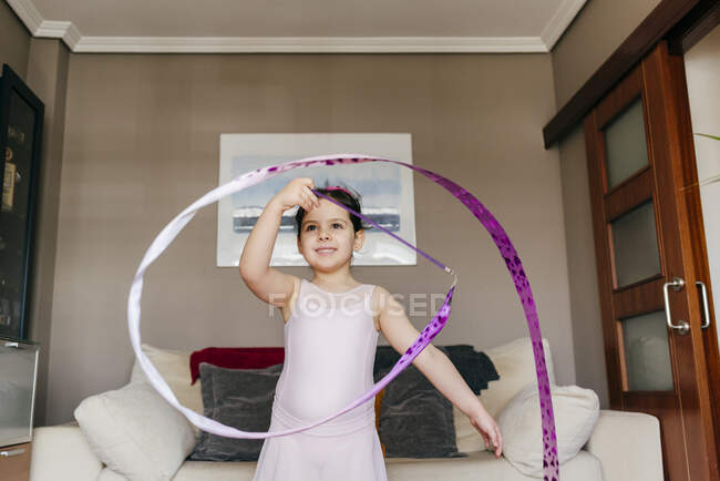Konzentriert glücklich süßes kleines brünettes Mädchen im Trikot beim Spinnen des Bandes beim rhythmischen Gymnastiktraining im gemütlichen Wohnzimmer zu Hause — Stockfoto