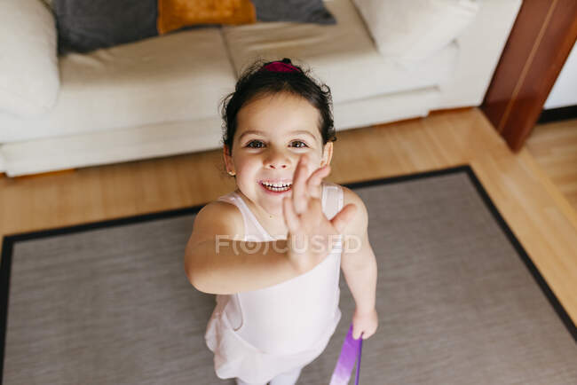 D'en haut petite fille avec ruban souriant pour la caméra et agitant la main pendant l'entraînement gymnastique rythmique à la maison — Photo de stock