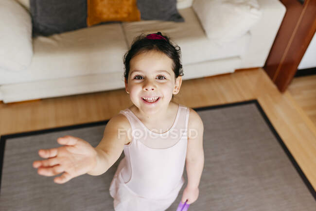 Сверху маленькая девочка с лентой, улыбающаяся перед камерой и машущая рукой во время гимнастических занятий дома — стоковое фото