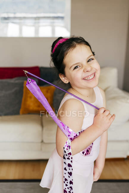 Visão lateral da linda menina morena feliz com fita sorrindo olhando para a câmera durante o treinamento de ginástica rítmica em casa — Fotografia de Stock