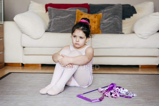 Sad little girl in leotard and tights embracing knees and sitting on floor near ribbon during unsuccessful gymnastic training in cozy living room at home — Stock Photo