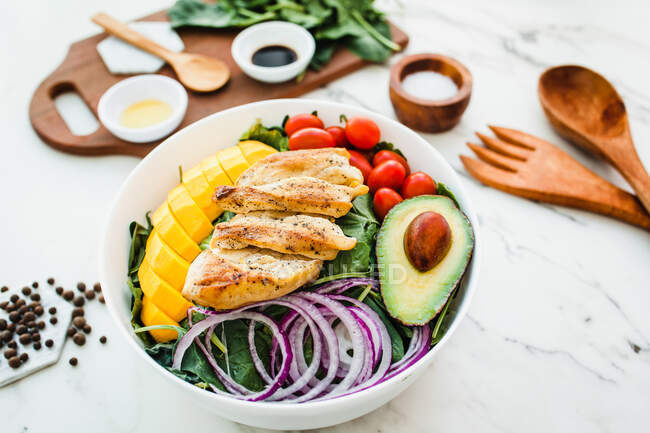 From above fried chicken and sliced mango placed near cherry tomatoes and half of avocado on spinach leaves and onion on marble table near utensils and sauces with spices — Stock Photo