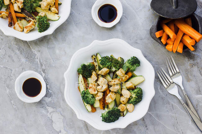 Von oben Blick auf Salat aus gebratenem Huhn und Zucchini mit Karotten und Brokkoli und in Schüssel in der Nähe von Sojasauce und Gabeln auf Marmor-Tischplatte platziert — Stockfoto