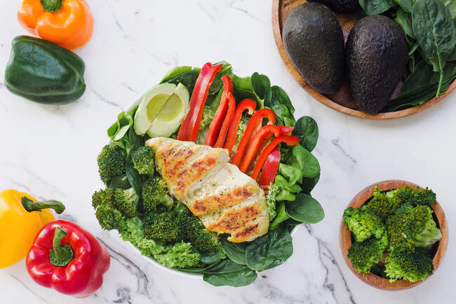Von oben gebratenes Huhn mit Brokkoli, Paprika und halbierter Avocado auf einem Bett aus grünen Blättern in einem Marmortisch — Stockfoto