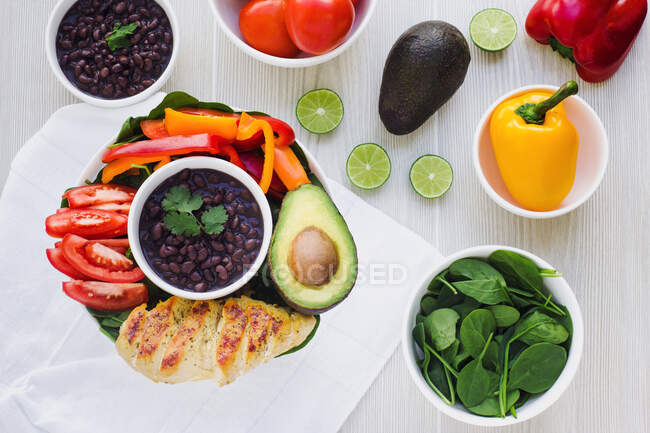 Fried chicken fillet and half of avocado placed in bowl near cut pepper and tomatoes around beans on napkin on table near slices of lime and spinach leaves — Stock Photo