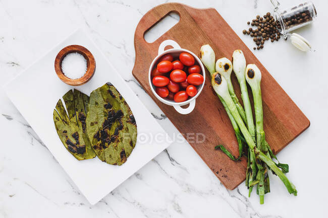 Top view of bowl with cherry tomatoes on wooden board with grilled green onion bulbs and seasoning on marble surface — Stock Photo