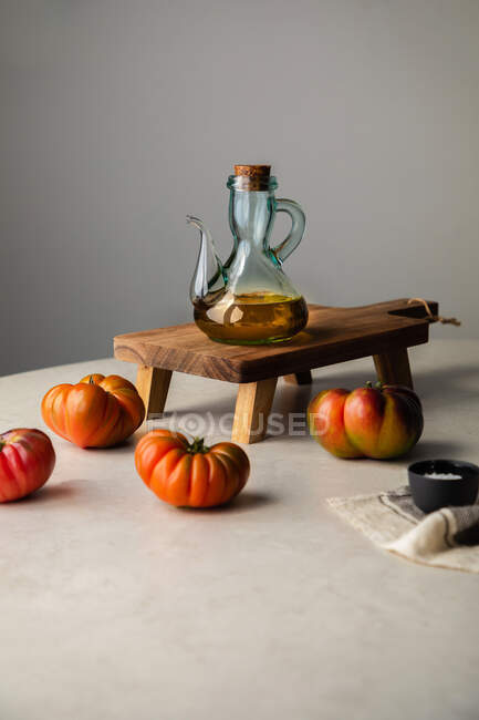 Tarro de vidrio con aceite de oliva colocado en un soporte de madera cerca de tomates rojos maduros frescos en la mesa de la cocina - foto de stock