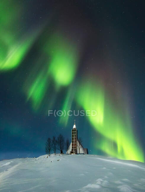 Amazing scenery of glowing Northern lights in dark sky over snowy terrain with small village church and trees in Iceland — Stock Photo