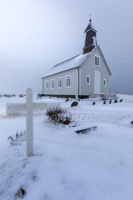 Низкий угол небольшой деревенской церкви, расположенной рядом с местным кладбищем с белым крестом среди снежного поля против серого пасмурного неба в зимний день в Исландии — стоковое фото