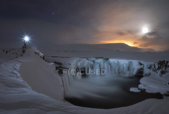 Unrecognizable traveler with glowing lantern standing on rock near small frozen lake with waterfalls, snowy terrain at sunset time in Iceland — Stock Photo