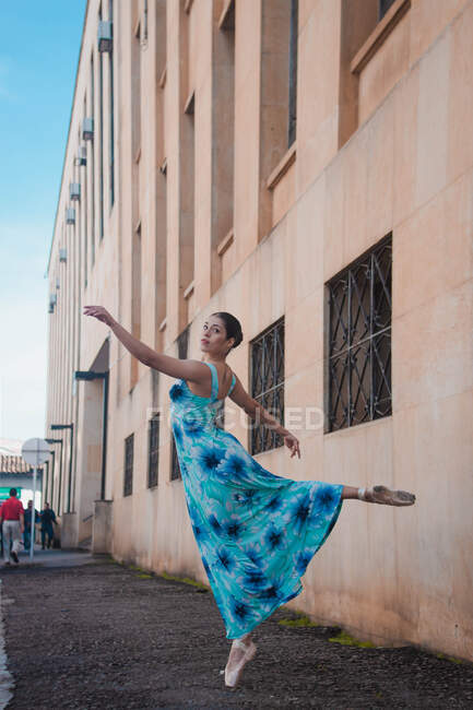 Vista lateral de la joven bailarina en vestido azul mirando a la cámara mientras baila con gracia fuera del edificio de mala calidad en la calle de la ciudad - foto de stock
