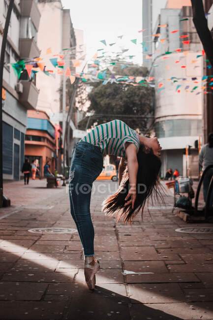 Mujer feliz en ropa casual haciendo curva trasera mientras baila en la calle en medio de edificios en la ciudad moderna - foto de stock