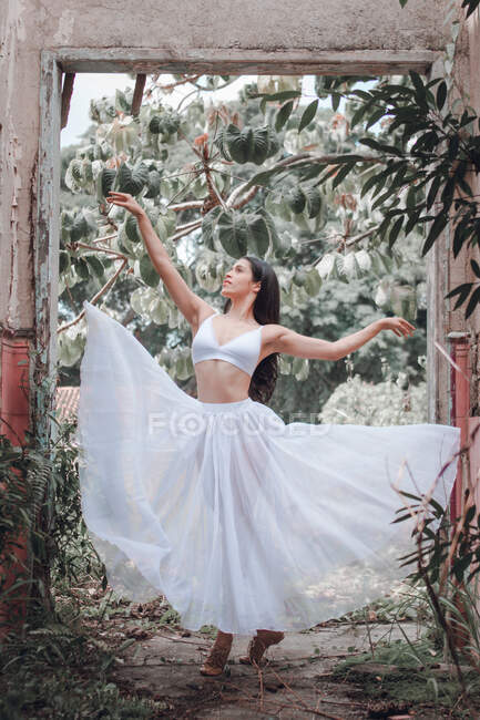 Full length young woman in white skirt and bra dancing with outstretched arms near green bushes and shabby arch in garden — Stock Photo