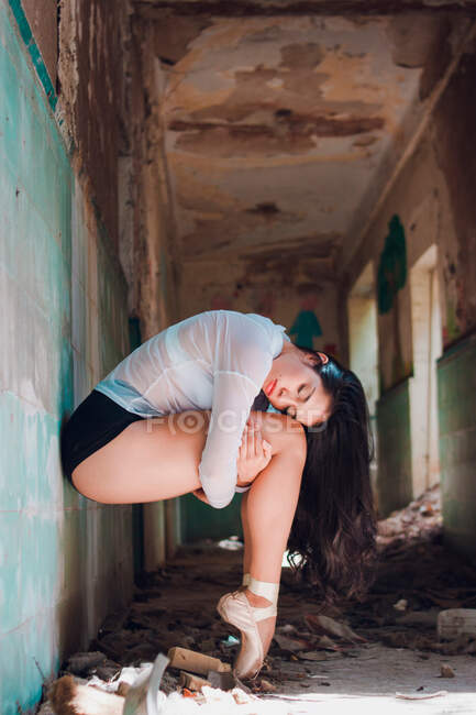 Seitenansicht einer schlanken Frau mit geschlossenen Augen, die Knie umarmt, während sie im schäbigen Flur eines verlassenen Gebäudes tanzt — Stockfoto
