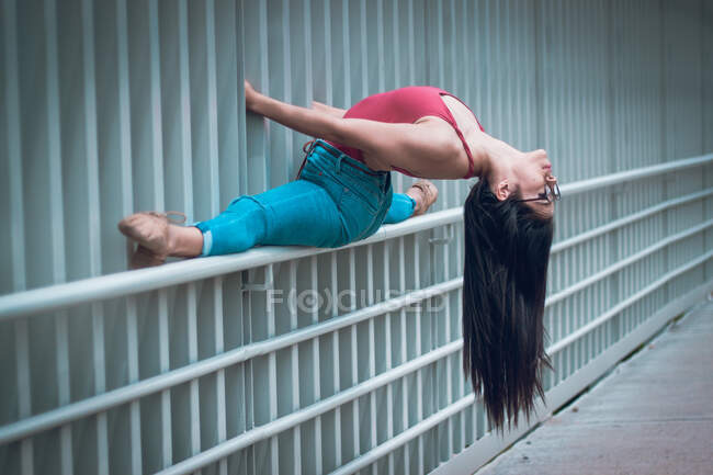 Side view of slip woman in casual clothes and glasses doing splits on fence and bending back while dancing on street — Stock Photo