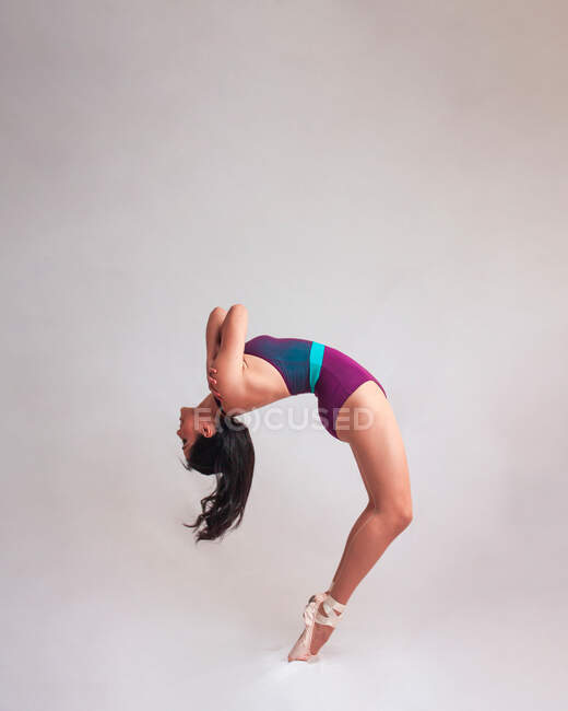 Side view of graceful woman in leotard and pointe shoes bending back while dancing against gray background — Stock Photo