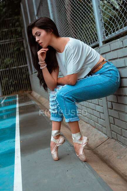 Vue latérale de la ballerine avec les yeux fermés dans des vêtements décontractés assis sur la pointe des pieds avec des chaussures de pointe sur une clôture murale en brique dans la rue — Photo de stock