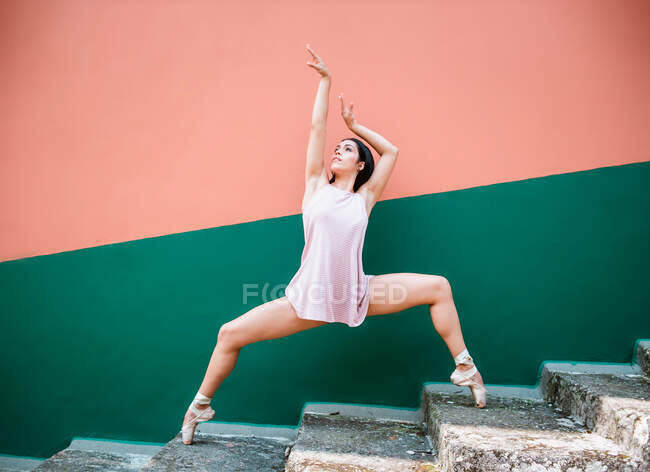 Brunette ballerina in pointe shoes standing on tip of toes on weathered steps and raising arms while dancing against striped wall on street — Stock Photo