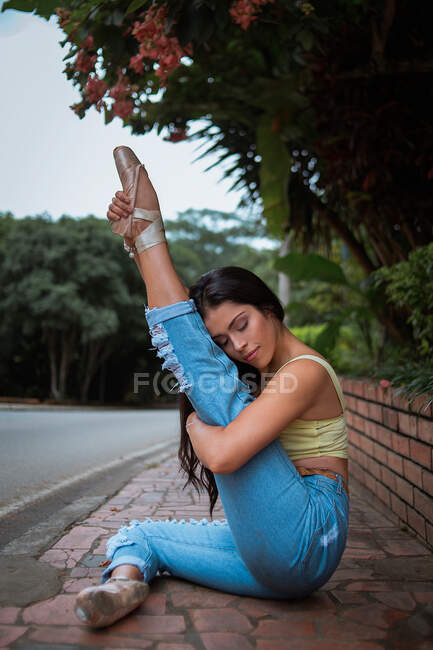 Seitenansicht einer schlanken Frau in lässiger Kleidung, die ihr Bein mit geschlossenen Augen hebt und streckt, während sie unter einem tropischen Baum am Straßenrand sitzt — Stockfoto