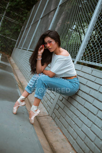 Vista lateral de la bailarina en ropa casual y gafas de sol sentadas apoyadas en puntillas con zapatos puntiagudos en la valla de pared de ladrillo en la calle - foto de stock