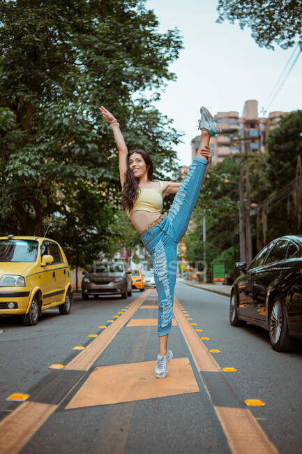 Donna in abiti casual facendo spacchi con braccio alzato e sorridente mentre balla su strada asfaltata in mezzo alle auto su strada trafficata della città moderna — Foto stock