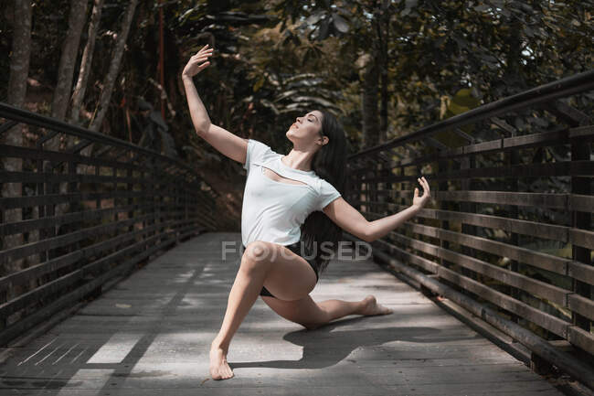 Barfüßige Frau mit ausgestreckten Armen und geschlossenen Augen tanzt an sonnigen Tagen auf Holzbrücke im friedlichen Garten — Stockfoto