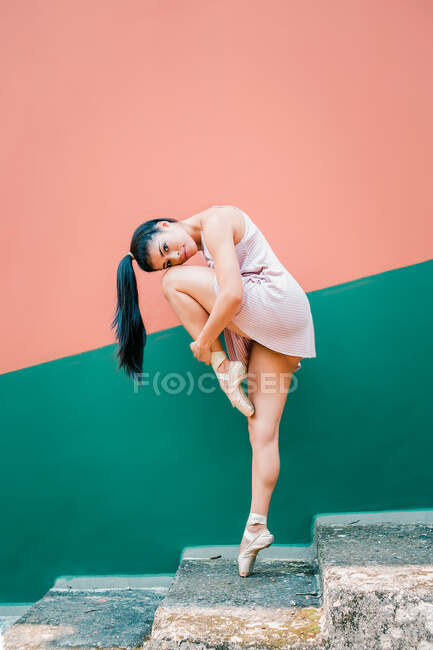 Ballerina looking at camera in pointe shoes standing on tip of toes embracing knees on weathered steps while dancing against striped colorful wall on street — Stock Photo