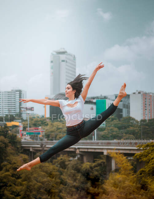 Босоніж жінка в повсякденному одязі стрибає і розпадається під час танців проти сучасного міста і хмарного неба — стокове фото