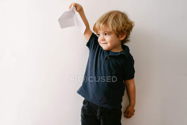 Bonito menino loiro em roupa casual brincando com o avião de papel sorrindo brilhantemente para a câmera no fundo da parede branca — Fotografia de Stock