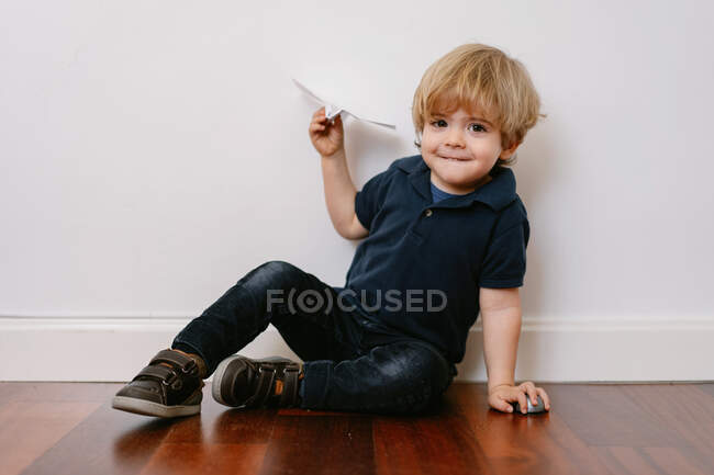 Lindo chico rubio en traje casual sentado en el suelo de madera jugando con el avión de papel sonriendo brillantemente a la cámara en el fondo blanco de la pared - foto de stock