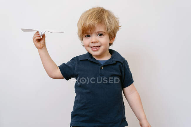 Cute blond boy in casual outfit playing with paper plane smiling brightly at camera on white wall background — Stock Photo