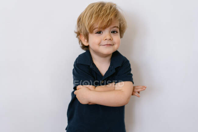 Чарівний хлопчик дошкільного віку в повсякденній футболці з руками схрестив усміхнений погляд на камеру, що спирається на білий фон стіни — стокове фото