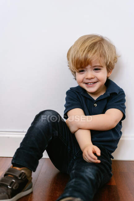 Очаровательный дошкольник в футболке, улыбаясь, смотрит в сторону, сидя на деревянном полу и наклоняясь на белом фоне стены — стоковое фото