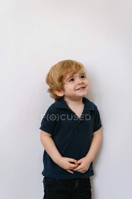 Adorable niño preescolar en camiseta casual sonriendo mirando hacia otro lado apoyado en un fondo blanco de la pared - foto de stock