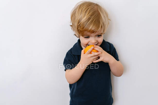 Petit garçon blond drôle en vêtements décontractés mangeant la moitié de l'orange en regardant loin avec le sourire tout en se tenant sur fond blanc — Photo de stock