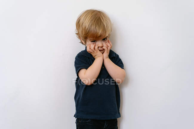 Annoyed blond little boy in casual clothes looking away with dissatisfaction while standing on white wall with hands on face — Stock Photo
