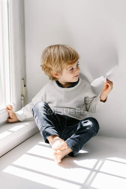 Joyeux petit enfant blond en vêtements décontractés jouant avec l'avion en papier tout en étant assis pieds nus sur le rebord de la fenêtre le jour ensoleillé — Photo de stock