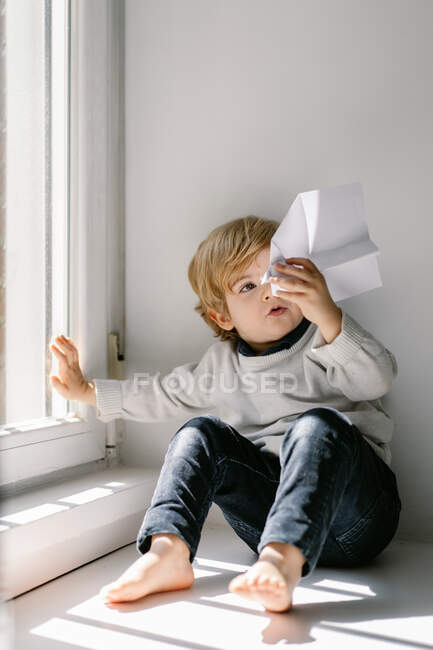 Glückliches blondes kleines Kind in lässiger Kleidung, das an sonnigen Tagen barfuß auf der Fensterbank sitzt und mit Papierflugzeug spielt — Stockfoto