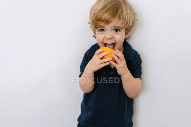 Lustiger blonder kleiner Junge in lässiger Kleidung, der halb orange isst, in die Kamera schaut und lächelnd die Zunge herausstreckt, während er vor weißem Hintergrund steht — Stockfoto