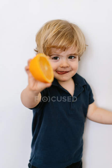Divertido niño rubio con ropa casual que se extiende ofreciendo la mitad de naranja hacia la cámara y sacando la lengua con sonrisa mientras está de pie contra el fondo blanco - foto de stock