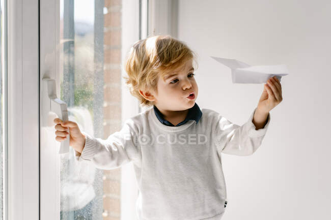 Glückliches blondes Kind in lässiger Kleidung, das an sonnigen Tagen barfuß auf der Fensterbank sitzt und mit Papierflugzeug spielt — Stockfoto