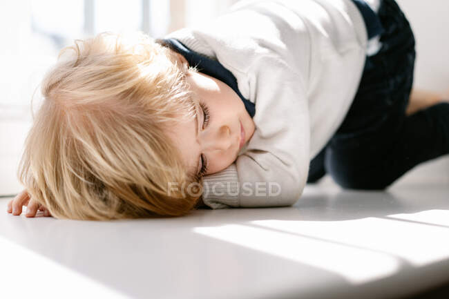 Vista lateral del tranquilo chico rubio en ropa casual durmiendo mientras descansa en el alféizar de la ventana en un apartamento moderno en un día soleado - foto de stock