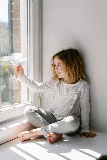Feliz niña rubia en pijama jugando con el avión de papel mientras está sentado descalzo en el alféizar de la ventana en el día soleado - foto de stock