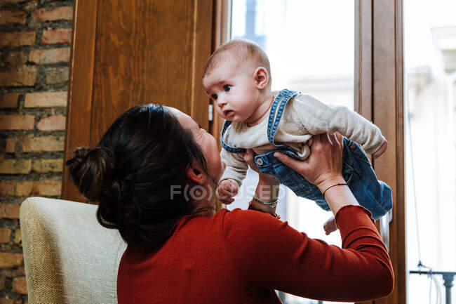 Entzückte Frau hebt süßes Baby und spielt auf Stuhl am Fenster in gemütlichem Zimmer zu Hause — Stockfoto