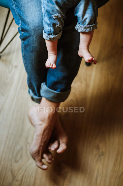 Свыше анонимный босой родитель в джинсах сидит на стуле с ребенком в уютной комнате дома — стоковое фото