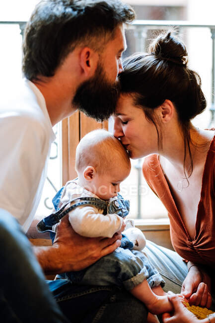 Glückliche Eltern küssen Baby und einander, während sie am Fenster im gemütlichen Zimmer am Wochenende zu Hause sitzen — Stockfoto